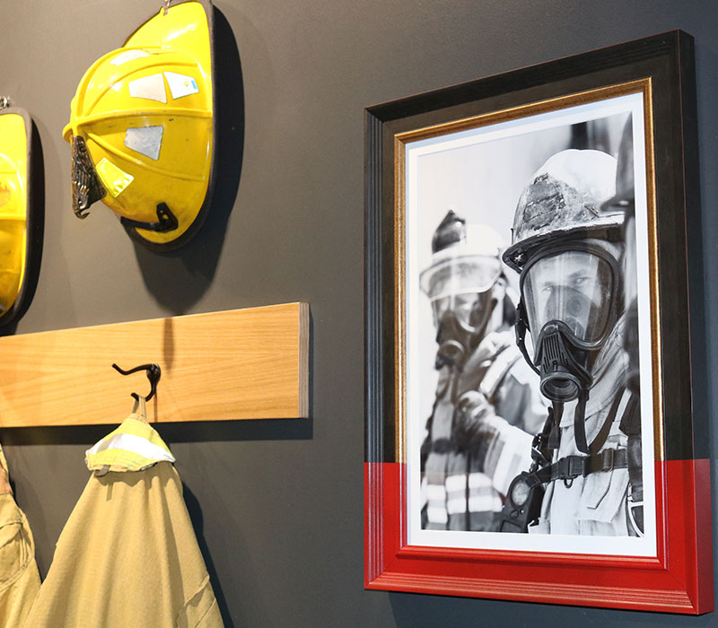 Wall display inside a Firehouse Subs restaurant featuring yellow firefighter helmets, a hanging firefighter jacket, and a framed black-and-white photo of firefighters in full gear.
